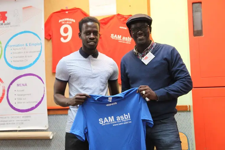 Deux hommes présentent un maillot de football floqué SAM asbl, devant les maillots des Diambars de Liège.