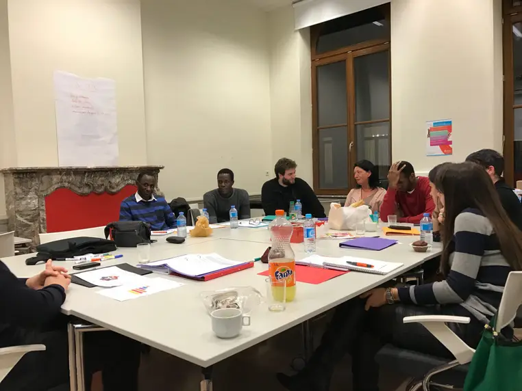 Une dizaine de personnes en réunion autour d'une grande table, un paperboard intitulé Actions au mur.