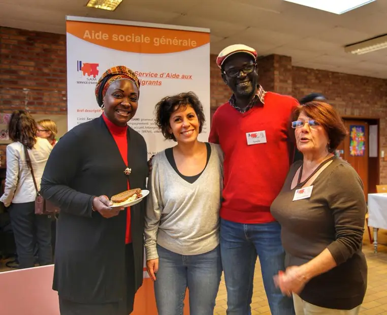 Quatre personnes discutent devant un panneau Aide sociale générale du Service d'Aide aux Migrants.