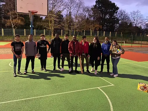 Un groupe de jeunes réunis sur un terrain multisports de plein air, ballon à la main.